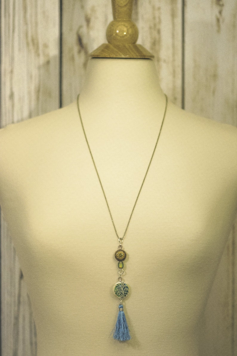 Puede incluir: Un collar largo de cadena plateada con un borla azul y tres colgantes redondos. Los colgantes est&aacute;n decorados con patrones verdes, marrones y dorados.