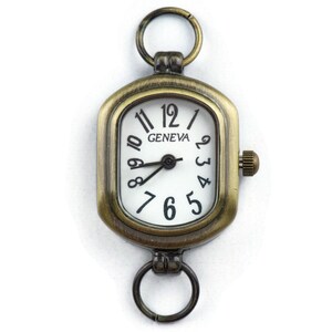 Pu&ograve; includere: Un quadrante di orologio di forma quadrata di colore oro antico con numeri romani neri e sfondo bianco. Il quadrante dell'orologio dice "GENEVA" nella parte superiore. L'orologio ha un anello nella parte superiore e inferiore per fissarlo a una catena o un braccialetto.