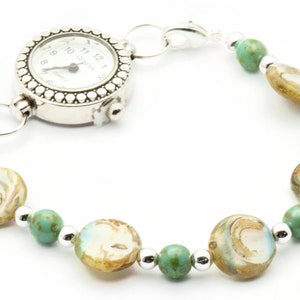 Op de afbeelding: Een zilveren zakhorloge met een witte wijzerplaat en zwarte wijzers is bevestigd aan een armband gemaakt van turquoise en bruine kralen.