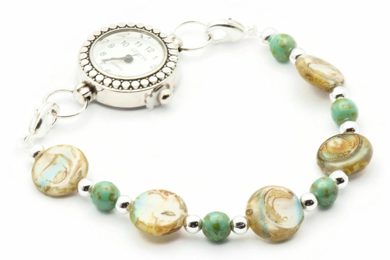 Op de afbeelding: Een zilveren zakhorloge met een witte wijzerplaat en zwarte wijzers is bevestigd aan een armband gemaakt van turquoise en bruine kralen.