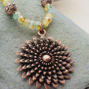 Pu&ograve; includere: Collana con pendente color rame con un design floreale dettagliato. Il pendente &egrave; sospeso a una catena di perline gialle, verdi e marroni. La collana &egrave; appoggiata su una superficie grigia e strutturata.