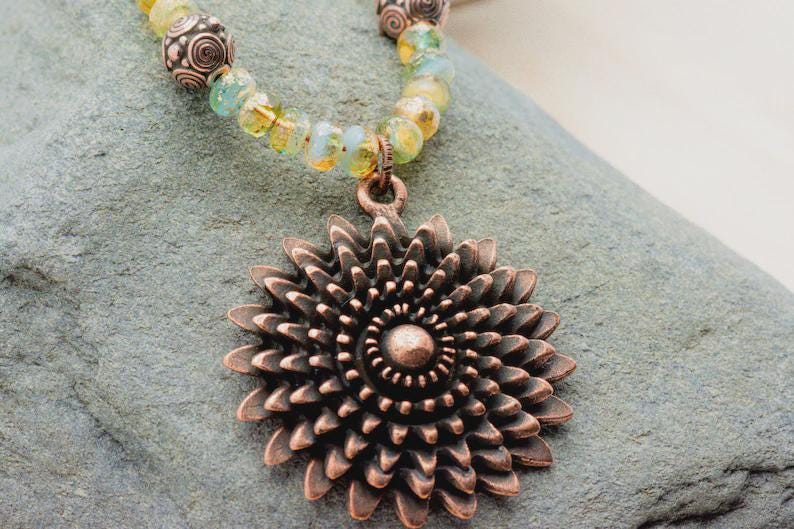 Pu&ograve; includere: Collana con pendente color rame con un design floreale dettagliato. Il pendente &egrave; sospeso a una catena di perline gialle, verdi e marroni. La collana &egrave; appoggiata su una superficie grigia e strutturata.