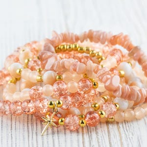 May include: A stack of bracelets made with peach-coloured sunstone and clear glass beads. The bracelets have gold accents and one bracelet has a gold star charm.