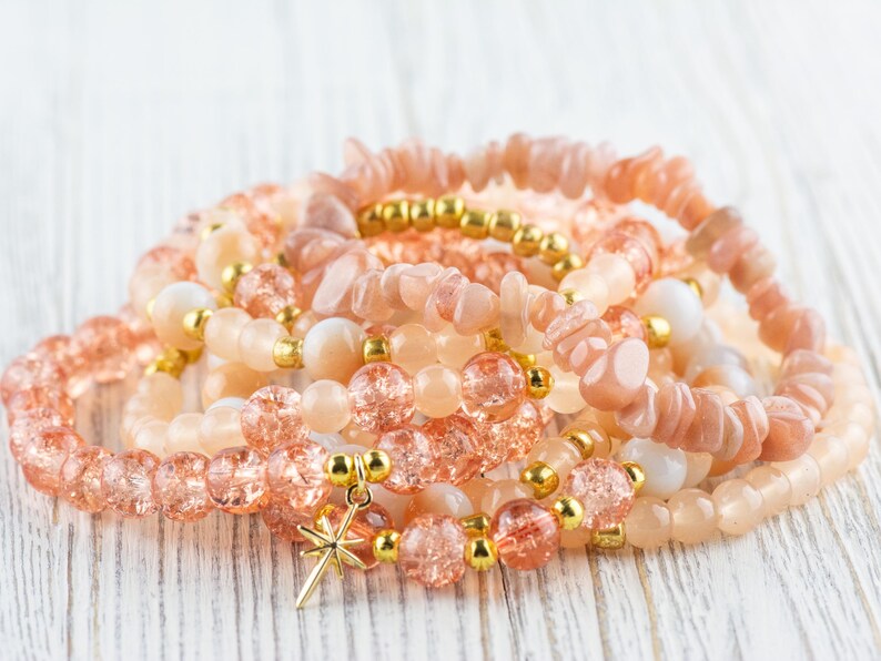 May include: A stack of bracelets made with peach-coloured sunstone and clear glass beads. The bracelets have gold accents and one bracelet has a gold star charm.