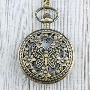 K&ouml;nnte beinhalten: Antike bronzefarbene Taschenuhr mit Schmetterlingsdesign. Das Zifferblatt ist durch ein dekoratives Blumen- und Schmetterlingsfiligran sichtbar. Die Uhr h&auml;ngt an einer passenden Kette, vor einem wei&szlig;en Holzhintergrund.