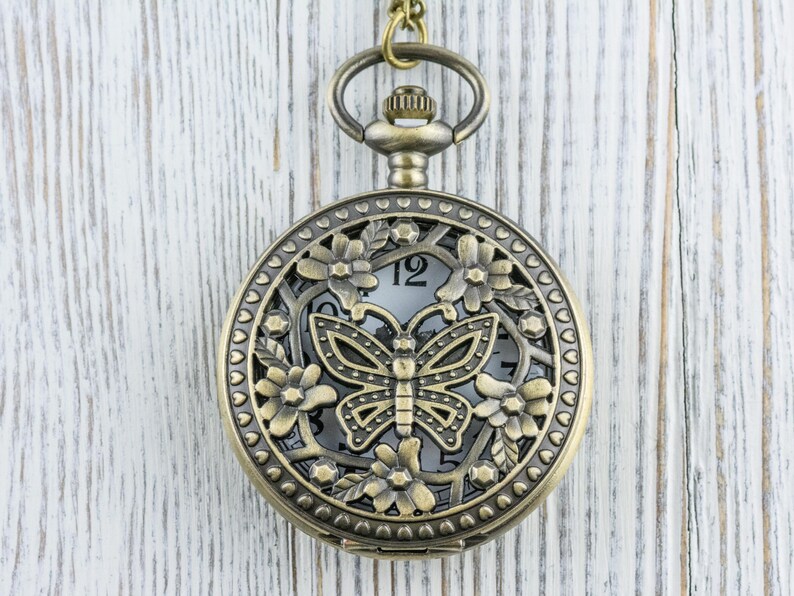 K&ouml;nnte beinhalten: Antike bronzefarbene Taschenuhr mit Schmetterlingsdesign. Das Zifferblatt ist durch ein dekoratives Blumen- und Schmetterlingsfiligran sichtbar. Die Uhr h&auml;ngt an einer passenden Kette, vor einem wei&szlig;en Holzhintergrund.
