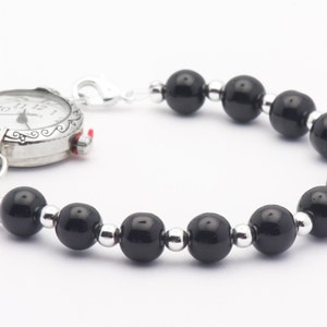 May include: A silver pocket watch with a black beaded bracelet. The watch face has black Roman numeral hour markers and black hands.