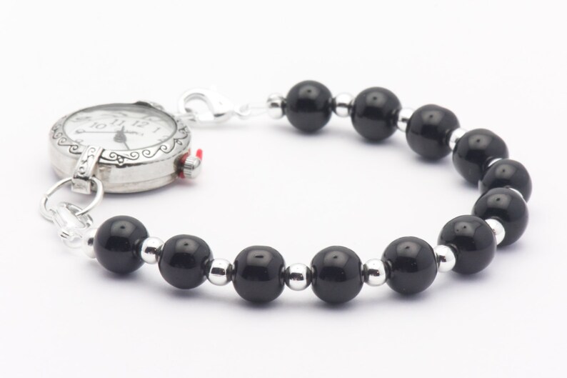 May include: A silver pocket watch with a black beaded bracelet. The watch face has black Roman numeral hour markers and black hands.