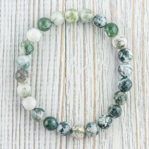 May include: A green and white agate stone bracelet with round beads. The bracelet is on a white wooden background.