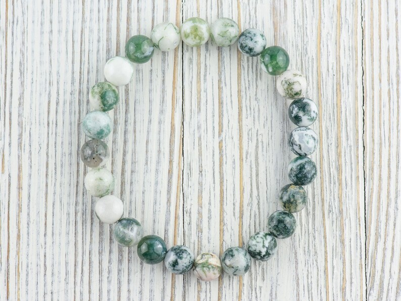 May include: A green and white agate stone bracelet with round beads. The bracelet is on a white wooden background.