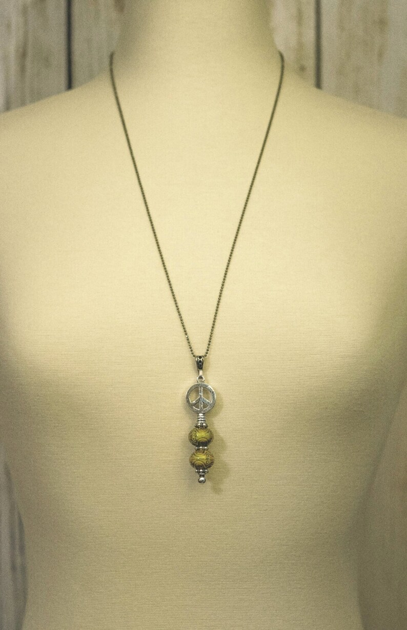Puede incluir: Un collar de cadena de plata con un dije de signo de paz y tres cuentas doradas.