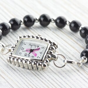 May include: A silver-toned watch with a square face and a pink ribbon design, attached to a bracelet of black, round beads. The watch face displays the time and the pink ribbon, a symbol of breast cancer awareness.