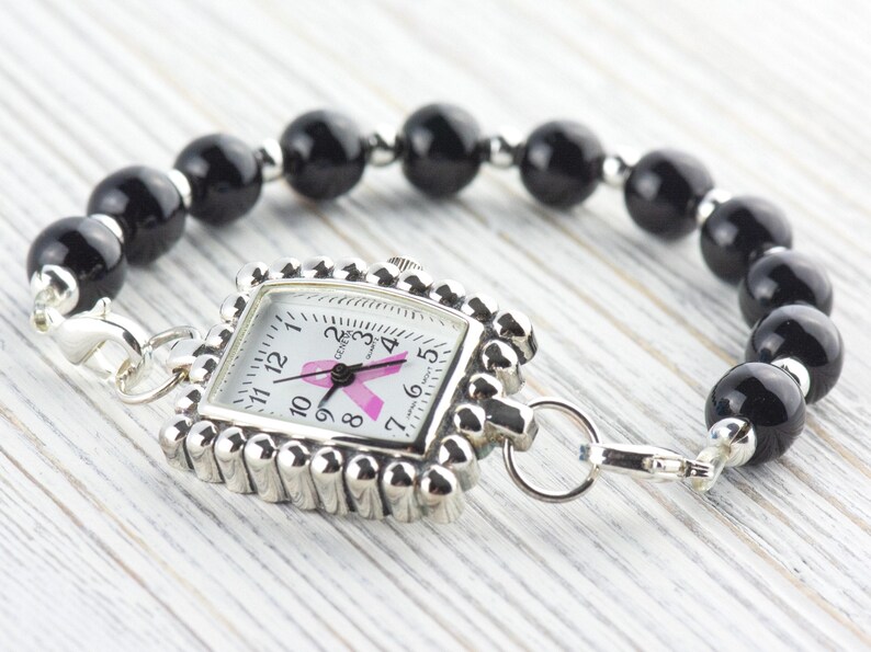 May include: A silver-toned watch with a square face and a pink ribbon design, attached to a bracelet of black, round beads. The watch face displays the time and the pink ribbon, a symbol of breast cancer awareness.