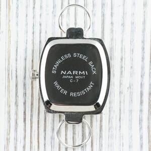 Puede incluir: Reloj plateado con una esfera cuadrada y una parte posterior de acero inoxidable. La esfera del reloj dice "NARMI JAPAN MOVT C-7 WATER RESISTANT".
