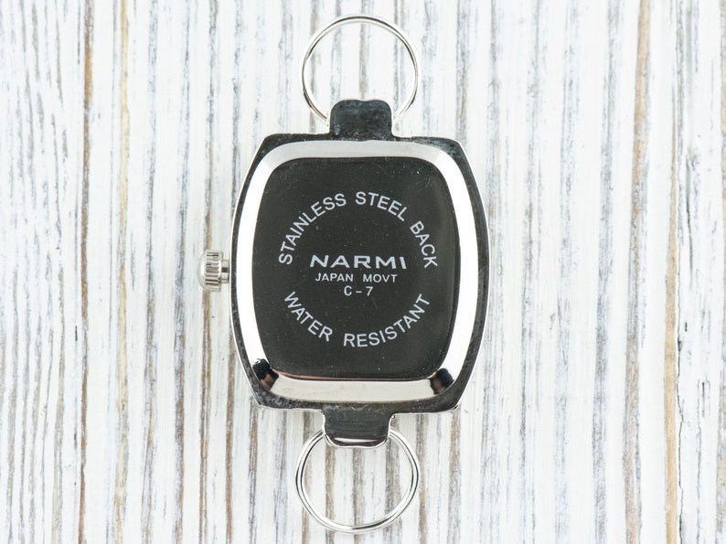 Puede incluir: Reloj plateado con una esfera cuadrada y una parte posterior de acero inoxidable. La esfera del reloj dice "NARMI JAPAN MOVT C-7 WATER RESISTANT".