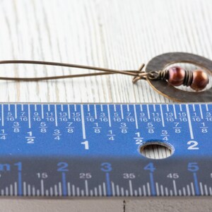 May include: A handcrafted bronze earring with a hammered oval pendant and two small, reddish-brown beads. The earring's total length is approximately 2 inches.