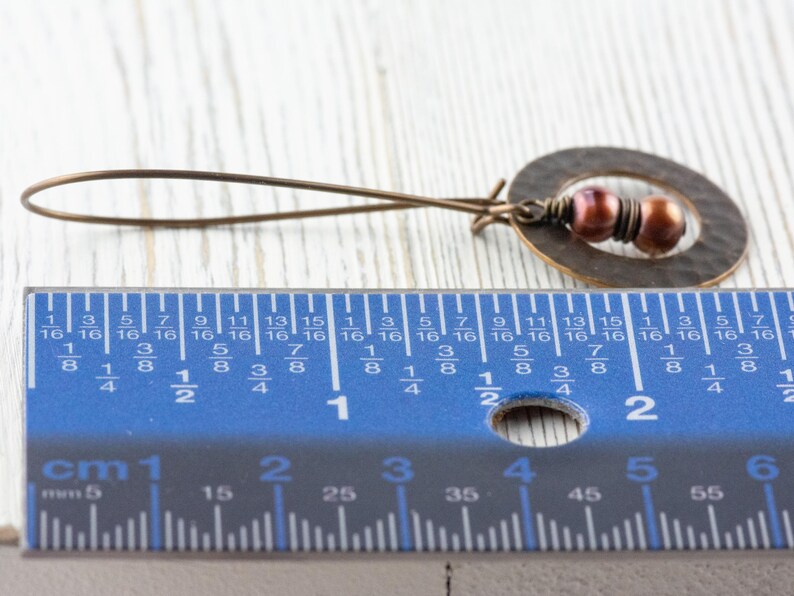 May include: A handcrafted bronze earring with a hammered oval pendant and two small, reddish-brown beads. The earring's total length is approximately 2 inches.