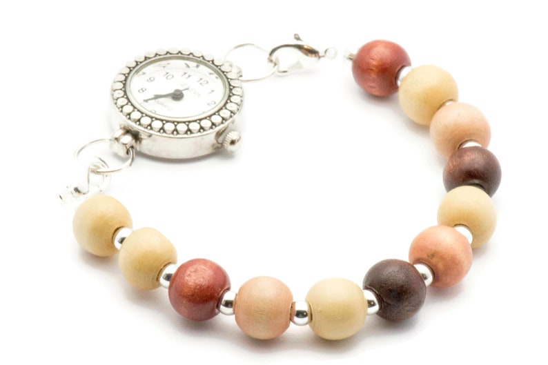Puede incluir: Un reloj plateado con una esfera redonda y un bisel de cuentas. El reloj est&aacute; unido a una pulsera hecha de cuentas de madera en tonos marrones, beige y rosa.