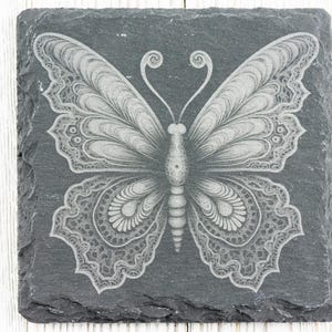 Puede incluir: Un posavasos de pizarra negra con un dise&ntilde;o de mariposa blanca intrincada grabado en la superficie.