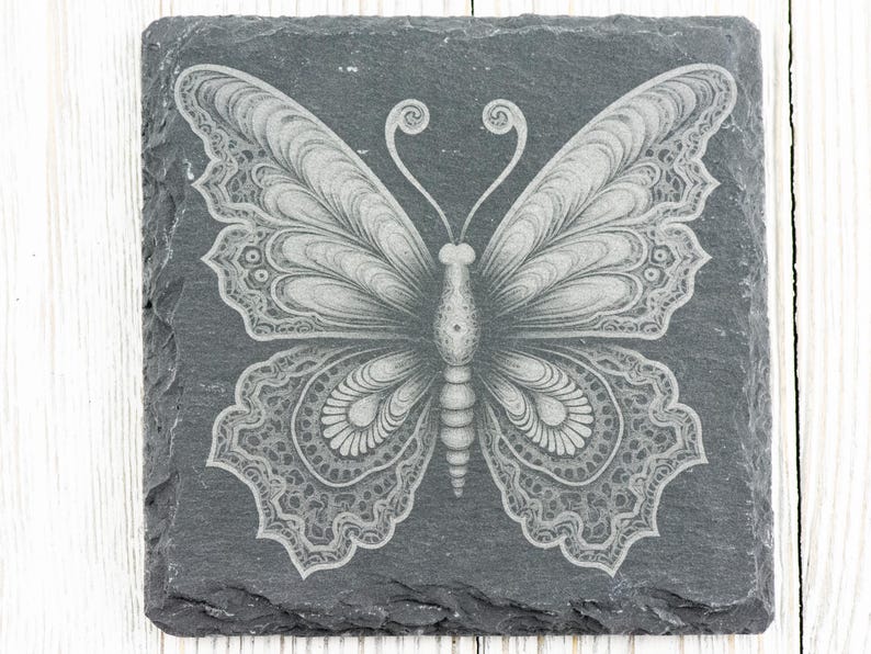 Puede incluir: Un posavasos de pizarra negra con un dise&ntilde;o de mariposa blanca intrincada grabado en la superficie.
