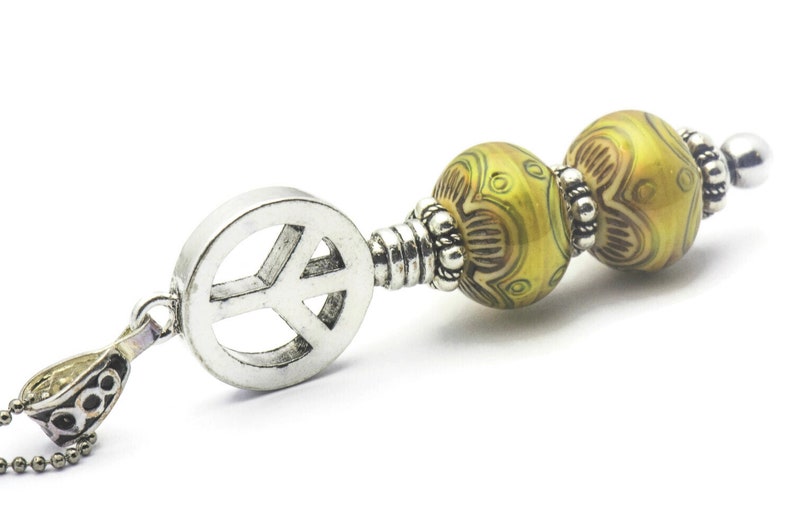 Puede incluir: Un collar con un dije de plata con el signo de la paz y dos cuentas de vidrio con patrones verdes y marrones.