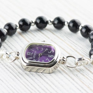 May include: A Geneva watch with a square silver face and a purple dial, connected to a bracelet of large, glossy black beads. The watch features silver clasps and is set against a light wood grain background.