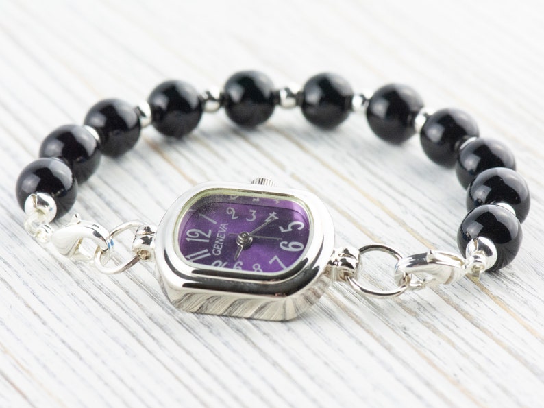 May include: A Geneva watch with a square silver face and a purple dial, connected to a bracelet of large, glossy black beads. The watch features silver clasps and is set against a light wood grain background.