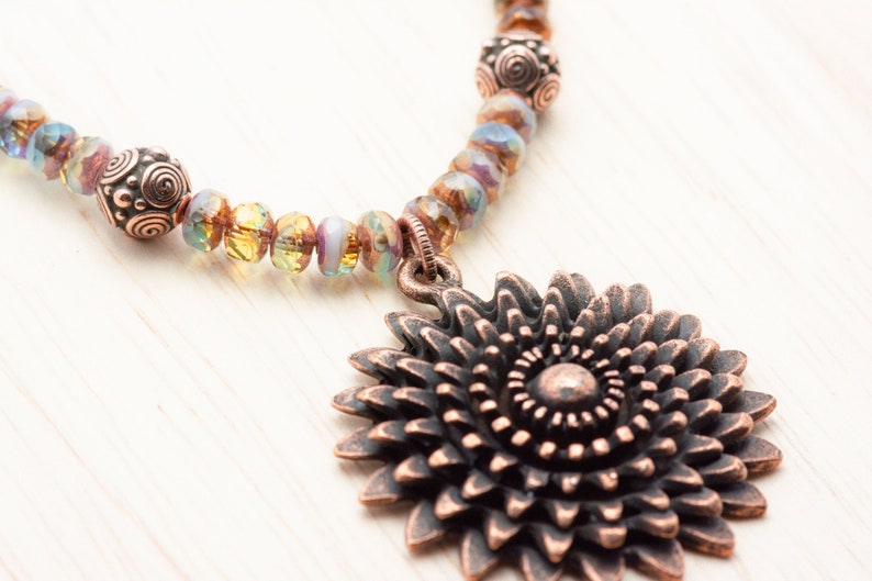 Puede incluir: Un collar de tono cobre con un gran colgante de girasol adornado. El collar est&aacute; hecho con cuentas de vidrio iridiscente y cuentas de tono cobre.