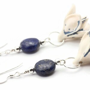 May include: A pair of earrings featuring blue lapis lazuli stones and white ceramic flower charms. The flowers have blue accents and are attached to silver hoops.
