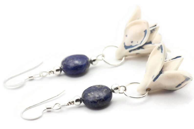 May include: A pair of earrings featuring blue lapis lazuli stones and white ceramic flower charms. The flowers have blue accents and are attached to silver hoops.
