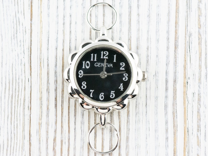 May include: A silver-toned watch with a black face and white numbers. The watch has a decorative scalloped edge and a chain with a ring at the top.