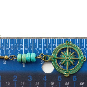 May include: A pair of turquoise and gold earrings with a compass charm. The earrings are approximately 3 cm long and feature a gold hook and a chain with turquoise beads.