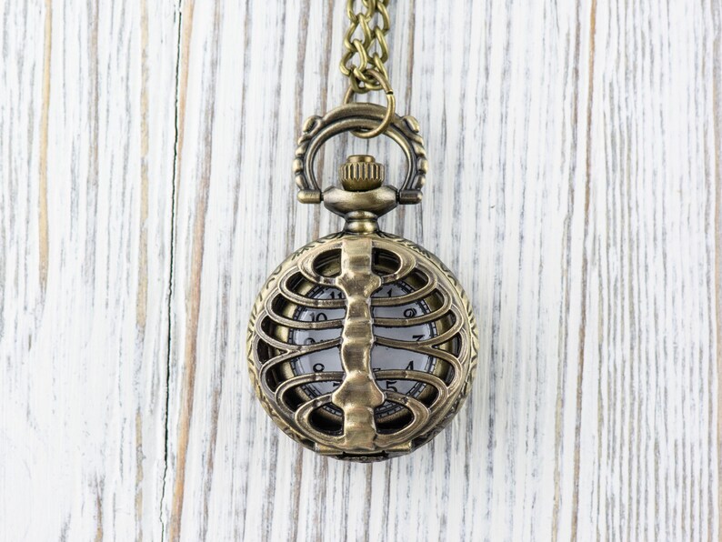 May include: A gold-toned pocket watch with a skeletonized design, featuring a ribcage pattern around the face. The watch is on a chain and hanging against a white wooden background.