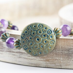 May include: A purple amethyst gemstone bracelet with a large, round, teal and gold patterned charm. The bracelet is on a white wooden surface.