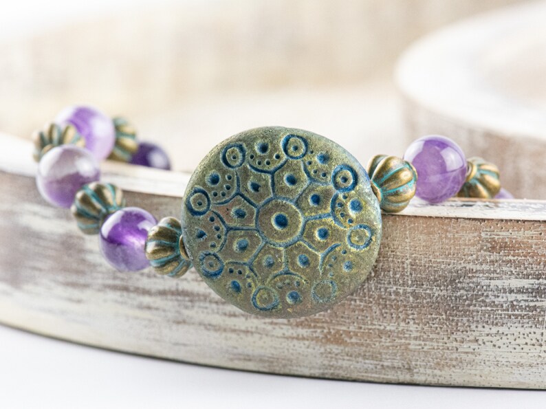 May include: A purple amethyst gemstone bracelet with a large, round, teal and gold patterned charm. The bracelet is on a white wooden surface.
