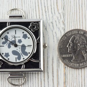 May include: A small, square, silver-toned watch with a black face and white hands. The watch face has a butterfly and star design. The watch is about the size of a US quarter.