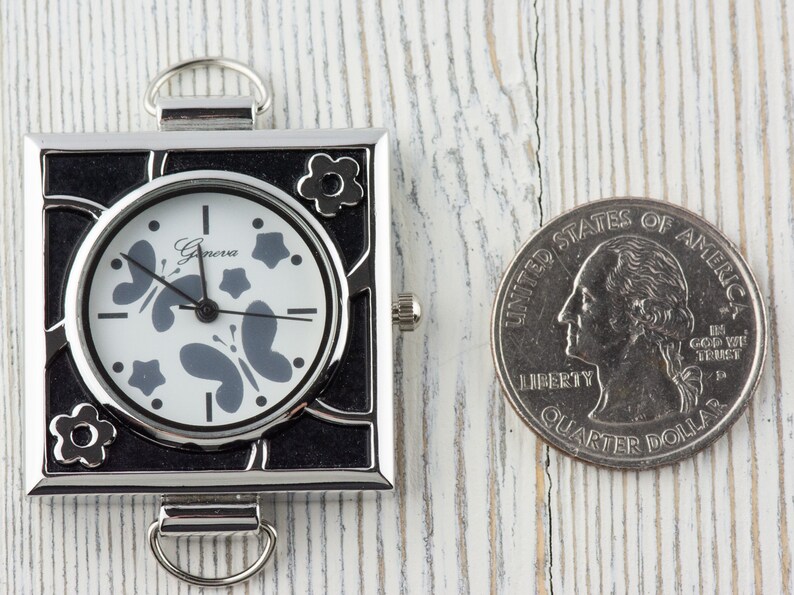 May include: A small, square, silver-toned watch with a black face and white hands. The watch face has a butterfly and star design. The watch is about the size of a US quarter.
