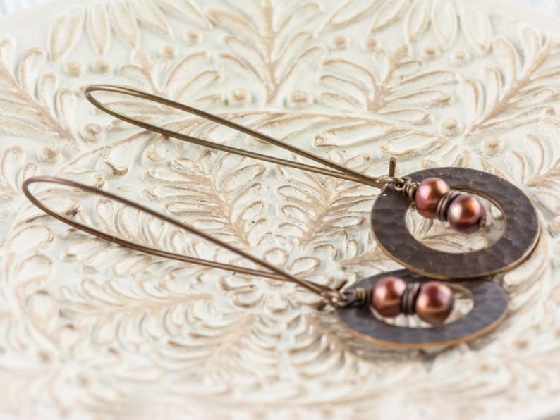 May include: A pair of handcrafted earrings featuring hammered copper oval and circular shapes. Each earring has two maroon-colored beads and long, thin copper wires for the ear. The earrings are displayed on a white, ornate surface.