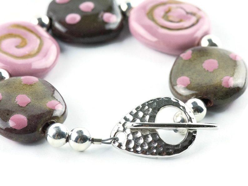 May include: A bracelet made of brown and pink ceramic beads with a silver clasp. The beads are decorated with pink polka dots and swirls.