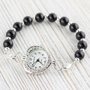 Pu&ograve; includere: Un orologio argentato a forma di cuore con un quadrante bianco e lancette nere. L'orologio &egrave; attaccato a un braccialetto di perline nere con una chiusura argentata.