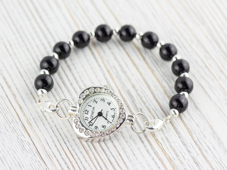 Pu&ograve; includere: Un orologio argentato a forma di cuore con un quadrante bianco e lancette nere. L'orologio &egrave; attaccato a un braccialetto di perline nere con una chiusura argentata.