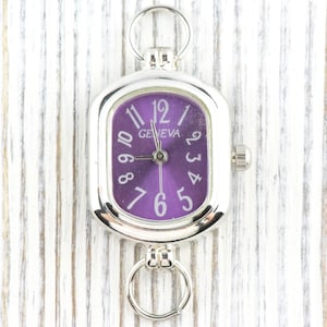 May include: Silver-toned Geneva watch with a purple face and white numerals. The watch has a rectangular face with rounded corners and a small crown on the side. It features a ring at the top and bottom for attachment.
