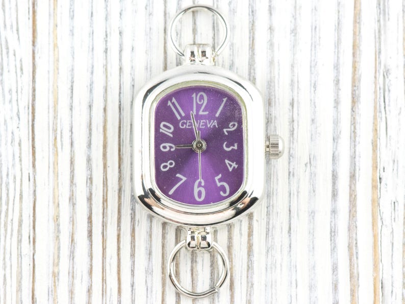 May include: Silver-toned Geneva watch with a purple face and white numerals. The watch has a rectangular face with rounded corners and a small crown on the side. It features a ring at the top and bottom for attachment.
