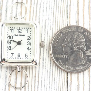 Puede incluir: Un reloj plateado con esfera cuadrada y blanca, con n&uacute;meros y manecillas negras. El reloj est&aacute; junto a una moneda de veinticinco centavos estadounidense. La esfera del reloj dice "NARMI".