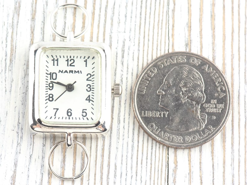 Puede incluir: Un reloj plateado con esfera cuadrada y blanca, con n&uacute;meros y manecillas negras. El reloj est&aacute; junto a una moneda de veinticinco centavos estadounidense. La esfera del reloj dice "NARMI".