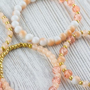 May include: Three beaded bracelets in shades of peach, white, and gold. The bracelets are made with different sized beads and are layered on top of each other.