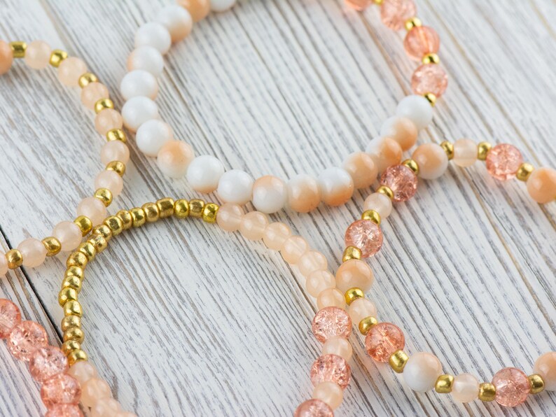 May include: Three beaded bracelets in shades of peach, white, and gold. The bracelets are made with different sized beads and are layered on top of each other.