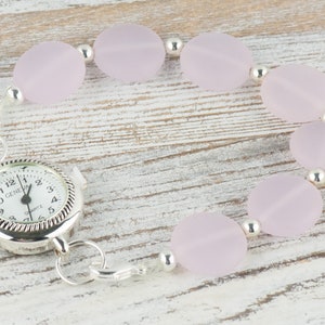 May include: A silver watch with a white face and black numbers is attached to a bracelet made of pale pink oval beads and silver spacers.