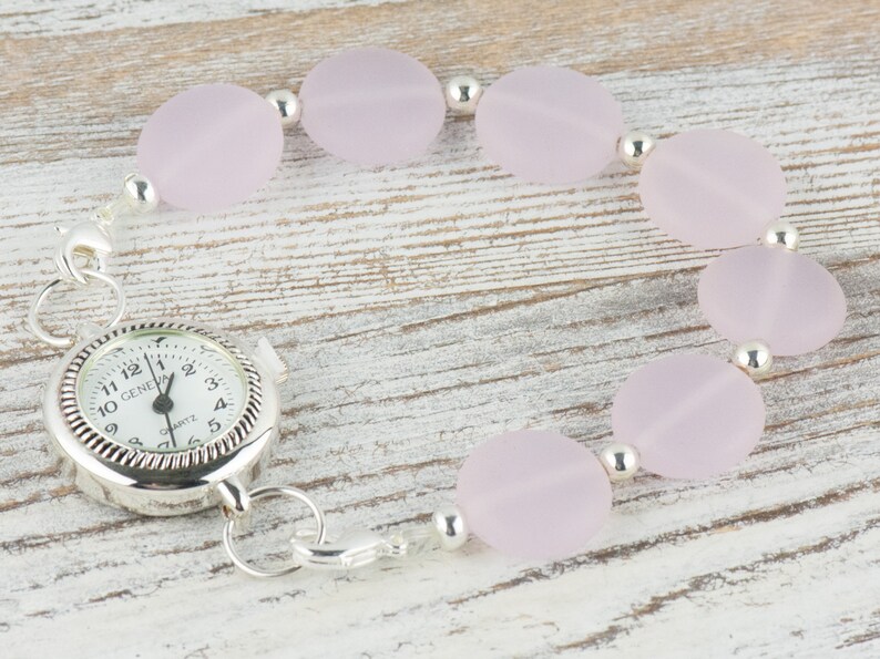 May include: A silver watch with a white face and black numbers is attached to a bracelet made of pale pink oval beads and silver spacers.