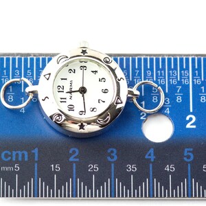 May include: A silver-tone round watch face with a white dial, black numerals, and the text NARMI. The bezel features engraved stars, moons, and spirals. Two silver rings are attached to the sides. A ruler shows the watch is 1 inch in diameter.